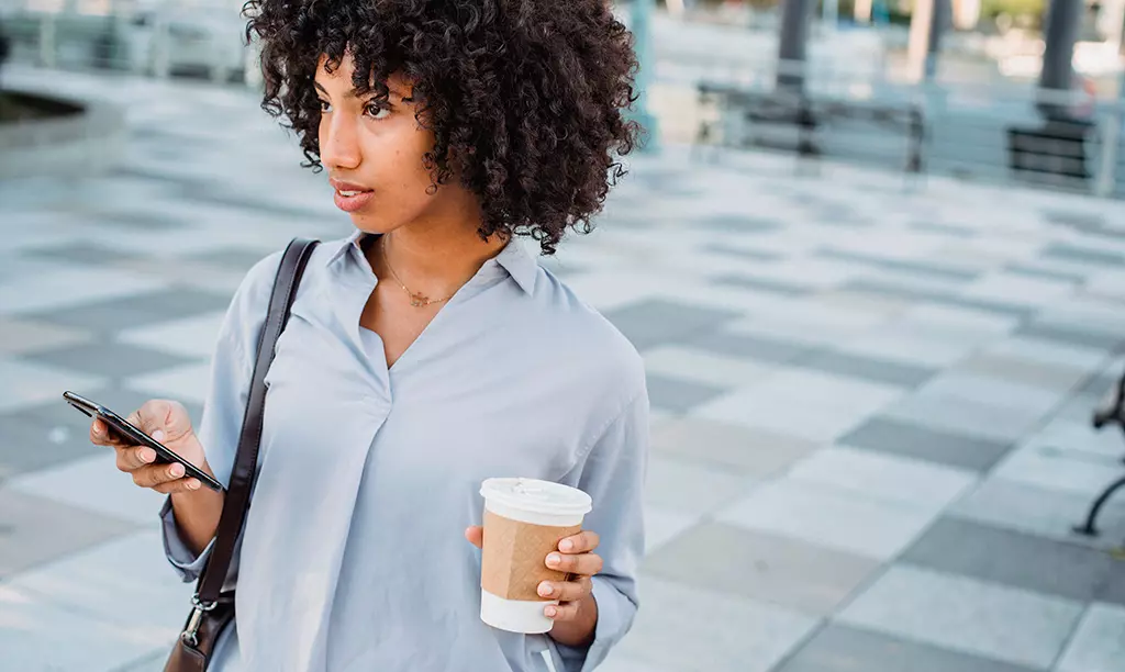 Pessoa caminhando ao ar livre segurando um smartphone e um copo de café em ambiente urbano moderno