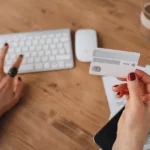 Pessoa segurando um cartão bancário enquanto utiliza um computador, com documentos financeiros sobre a mesa em ambiente de trabalho doméstico.