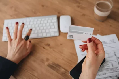 Pessoa segurando um cartão bancário enquanto utiliza um computador, com documentos financeiros sobre a mesa em ambiente de trabalho doméstico.