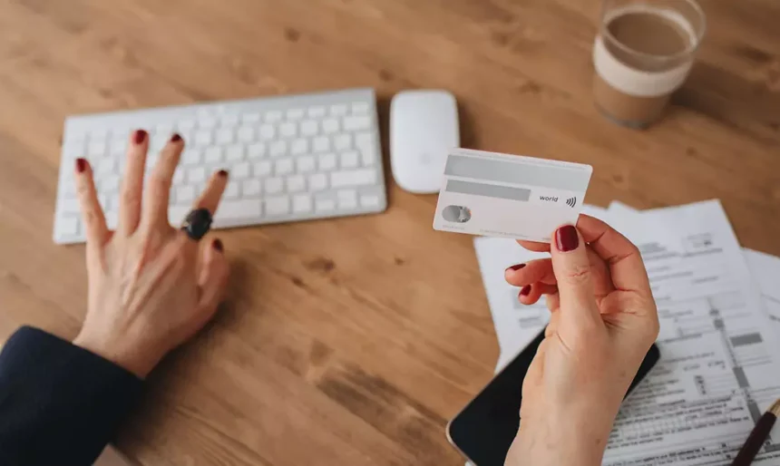Pessoa segurando um cartão bancário enquanto utiliza um computador, com documentos financeiros sobre a mesa em ambiente de trabalho doméstico.