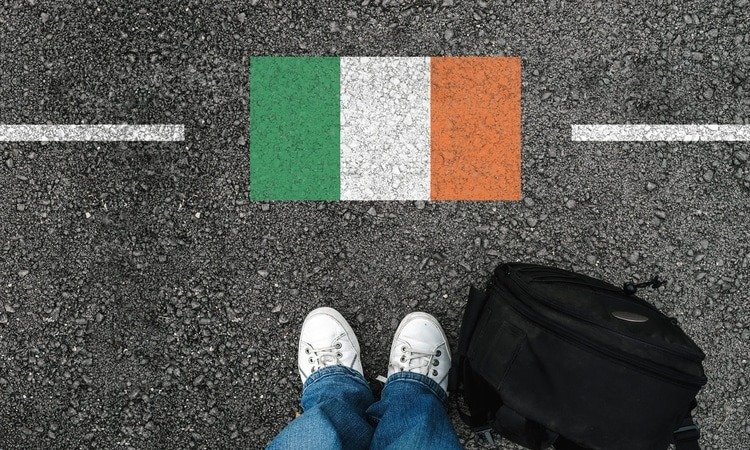 Pessoa com mala em frente à bandeira da Irlanda pintada no asfalto, representando imigração e mudança para o país.